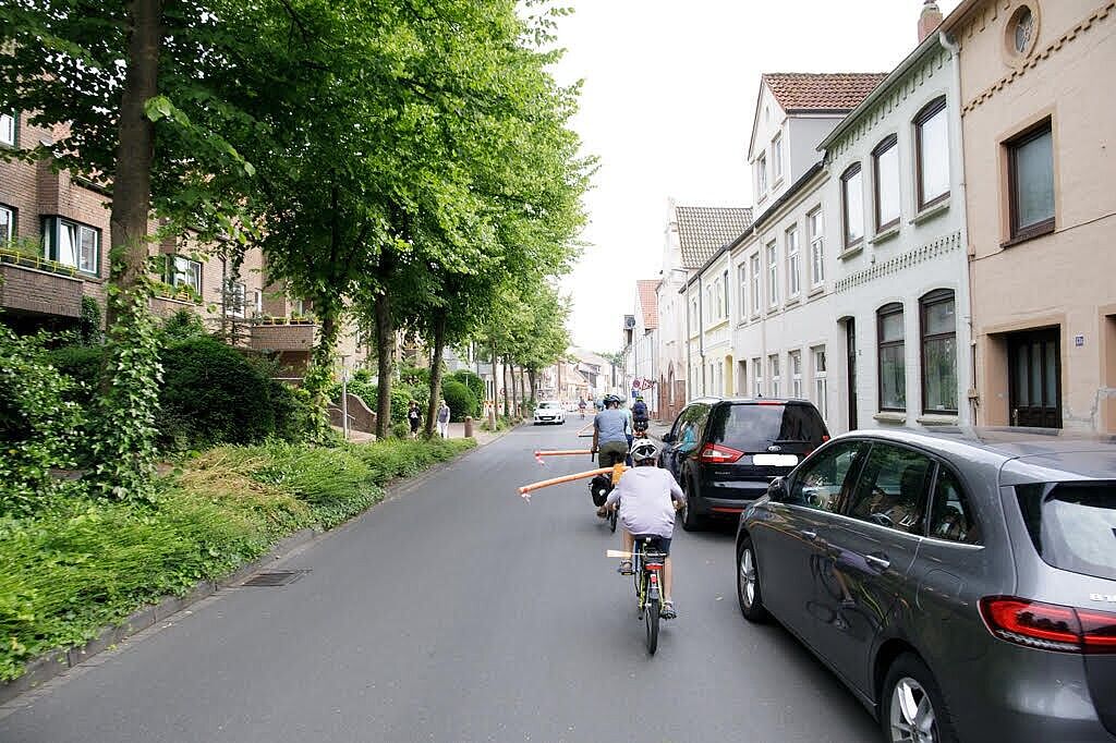 Fahrradfahrer mit Poolnudel am Gepäckträger fahren auf der Straße an parkenden Fahrzeugen vorbei.